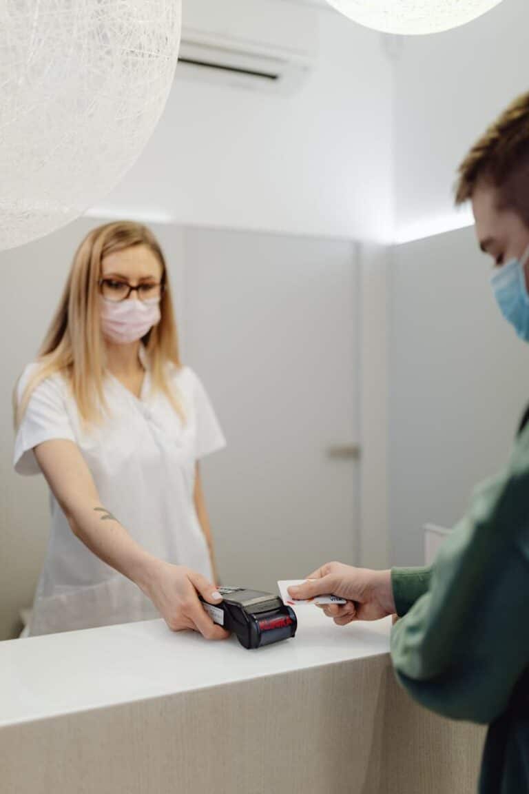 A masked patient pays at a clinic counter, using a card terminal handled by a healthcare professional.
